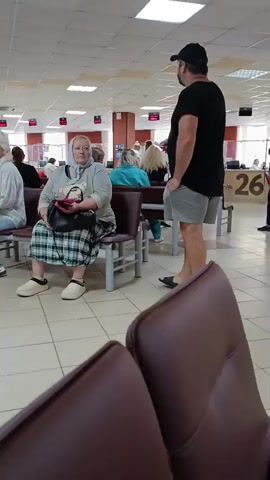 The video shows the interior of a building, likely a government office or public service center. Several people are present, seated and standing. In the foreground, a dark brown chair is visible. Further back, a woman wearing a headscarf and a plaid skirt sits on a chair, holding a black purse. A man in a black t-shirt and grey shorts stands nearby, facing away from the camera. In the background, other individuals are seated, and some are interacting with what appear to be service windows or desks. Digital displays with numbers are visible on the walls, suggesting a queue or waiting system. The environment is brightly lit with fluorescent ceiling lights, and the floor is tiled. The overall impression is of a busy waiting area.