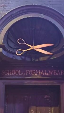 The video shows a close-up of the entrance to a shop called "School & Formalwear". Above the door, inside an arched, dark wooden frame, hangs a large, antique-looking pair of scissors. The scissors are rusty and appear to be made of metal. The inscription "SCHOOL & FORMALWEAR" is painted in red on the wooden architrave above the door. The door itself is dark wood and appears old. The brick wall of the building is visible in the background.