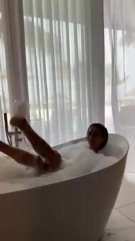 A woman is lying in a freestanding, white bathtub filled to the brim with bubbles. Her head and torso are visible as she smiles toward the camera. Another person’s legs are visible near the camera, with feet submerged in the bubbles. The bathtub is positioned in front of a large window covered with sheer white curtains. The view through the window reveals a beach scene. A modern-looking faucet is situated at one end of the bathtub.