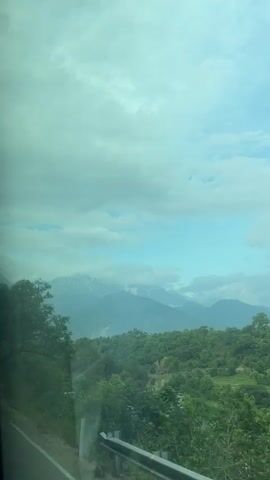 The video shows a view from inside a moving vehicle, likely a bus, looking out the window at a mountainous landscape. The sky is overcast with clouds, and the tops of the mountains are capped with snow. Lush green trees and vegetation cover the hillsides. A road is visible along the edge of the frame, and some blue structures, possibly greenhouses or shelters, are seen in the valley below. The camera moves as the vehicle travels along the road, passing by trees lining the roadside.