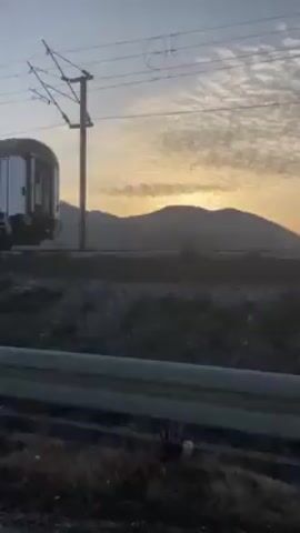 The video shows a passenger train passing by on a track. The train cars are covered in graffiti. The surrounding environment appears to be a roadside with gravel and sparse vegetation. A mountain is visible in the distance, silhouetted against a sunrise or sunset. The camera is moving, likely from inside a vehicle. Overhead electrical lines are visible.