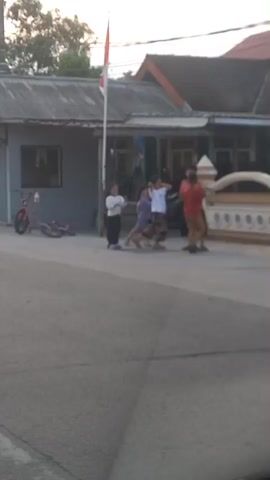 The video shows several children standing on a paved road in front of houses. A flag is flying on a pole. One child is on a bicycle and is riding past the group. Two other children are on bicycles lying on the ground to the left. The children are dressed in casual clothing. The scene appears to be a residential street.