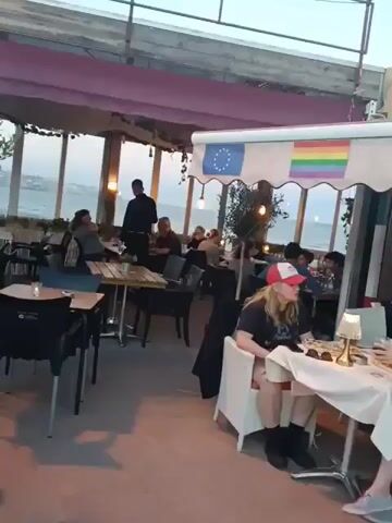 The video depicts the outdoor seating area of a restaurant or cafe. Several tables and chairs are arranged under a shaded awning, with small olive trees adding to the ambiance. Customers are seated at some of the tables, enjoying their meals or drinks. A waiter is seen serving food to a table. In the background, there is a glimpse of the beach and ocean. The overall atmosphere seems relaxed and pleasant.
