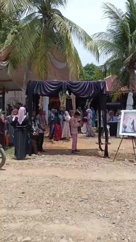 The video shows an outdoor gathering with many people. In the foreground, a motorcycle is parked. Behind it, a black draped archway stands over a pathway. People are standing and walking around the archway and in front of a building with a covered porch. Some people are wearing traditional Islamic clothing, including headscarves. There is a framed picture on an easel to the right of the archway. Palm trees and other foliage are visible in the background, suggesting a tropical or subtropical environment. The ground is covered in gravel and dirt. The scene appears to be a ceremony or celebration.