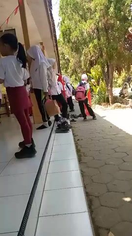 A group of young children, some wearing hijabs and school uniforms, are seen gathered in an outdoor walkway. The children are standing on a tiled floor, and a fence or railing runs along the edge of the walkway. To the right of the walkway, there is a path with loose stones and vegetation. The children appear to be waiting or engaged in some activity. One child in the foreground is wearing a white t-shirt and a maroon skirt over leggings. Other children are dressed in similar attire, with some carrying backpacks. The walkway is shaded by the roof of a building, and the background is filled with trees and greenery. Red and white flags are strung along the top of the walkway. The scene suggests a school or an outdoor gathering.