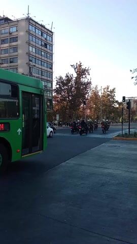 The video opens on a bright, sunny street corner in a city. A green and white bus waits on the side of the road, its door open. A motorcade of police motorcycles passes by in the distance.  People begin to cross the street, walking toward the camera. Most seem casual, with one man carrying a water bottle and another a potted plant. There is a small car waiting at the intersection to turn left as the light turns green. A tall, green-covered hill can be seen in the background.  A Chilean flag waves from the hill.