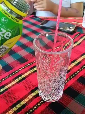 A bottle of Fuze Tea Lemon Lemongrass is being poured into a clear glass with a pink straw. The bottle is green with a yellow cap and the label indicates "Low Calories". The glass is ornately patterned and sits on a red, green, and yellow checkered tablecloth. The background shows a person's hands holding the bottle and another person's hands holding a phone. In the background, there are other glasses and a person sitting at the table. The lighting is natural and appears to be outdoors.