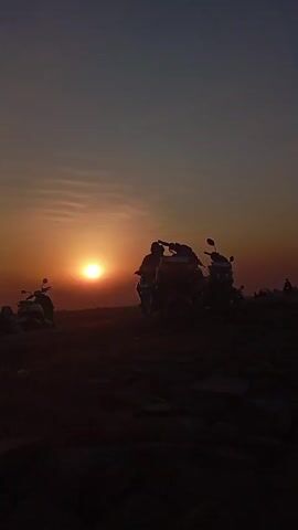 This is a vertical video of a sunset on a rocky incline. In the foreground, there are several large rocks. In the middle ground, there are two motorcycles parked side-by-side, with a third motorcycle to the left and slightly behind them. There are also two people on a motorcycle to the far left, silhouetted against the sun. The sun is low in the sky, casting a warm glow. In the background, there are a few other people standing or walking. The sky is a gradient of orange, pink, and purple.