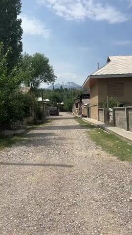 A gravel road runs through a village. Trees line the left side of the road, and buildings made of brick and mud are on both sides. A military truck is parked on the road, facing away from the camera. In the distance, snow-capped mountains rise above a treeline. The sky is blue with scattered clouds. The video is shot from a slightly elevated perspective, looking down the road towards the mountains. The lighting suggests it is daytime, likely late morning or early afternoon.