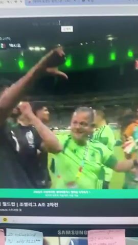 A computer monitor displays a soccer match replay. The screen shows a close-up of several male soccer players wearing green and black uniforms, some with a Mexican national team logo. They appear to be celebrating a victory, with some players raising their arms in the air and one player drinking from a water bottle. The score of the match is displayed on screen as 1-0. The background shows a stadium with green lighting. The lower part of the screen shows Korean text that appears to be information about the match, including the league name and match date. There are also several sticky notes attached to the monitor frame with handwritten text and IDs.