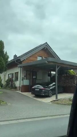 This media file captures a drive through a residential area in Braunfels, Germany, under a cloudy sky. The perspective is from inside a car, looking out the window.

The first scene shows a modern carport with a dark-colored car parked underneath. A large pile of neatly stacked firewood is a prominent feature next to the carport. Beyond the carport, a house with a sloping roof and a metal fence is visible. The yard appears somewhat unkempt, with bushes and some construction materials present.

As the drive continues, a white house with a chimney and a television antenna on the roof comes into view, surrounded by dense green trees. The next shot offers a close-up of the car's interior, showing the seatbelt and the edge of the passenger seat. The final scene presents another house with a white facade, partially obscured by lush green hedges.

The weather is overcast, suggesting it might be daytime, possibly late morning or afternoon. The overall impression is one of a suburban drive, with glimpses of everyday life in a German town. There is no visible text in the media file.