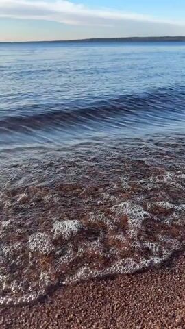 The video shows a calm body of water meeting a shoreline covered in small, dark-colored rocks. Gentle waves roll in and break on the shore, creating a shimmering effect on the wet rocks. In the distance, a treelined land mass is visible under a blue sky with wispy clouds. The scene appears tranquil and still.