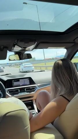 The video shows the interior of a car from a passenger's perspective, looking forward through the windshield. A woman with blonde hair is seated in the passenger seat, facing forward. The dashboard displays a navigation system. The car is on a highway, with other vehicles visible. The sky is clear and sunny, with a sunroof open. The audio features a song with lyrics.