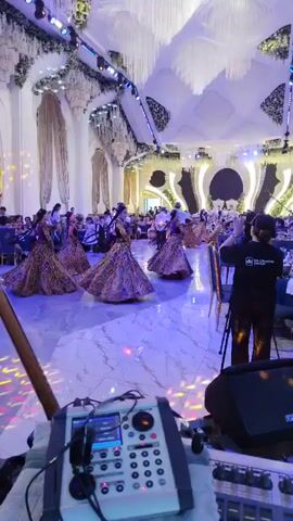 This video captures a lively event in a large, ornate hall, likely a wedding reception or similar celebration. In the foreground, a DJ setup is visible, with audio equipment, including mixers and speakers, arranged on a table. A guitar neck is also positioned at the lower left of the frame. The background features a spacious dance floor illuminated by colorful lights, with a group of dancers in traditional attire, characterized by their long, patterned skirts and dark tops. They are engaged in synchronized movements, with several of them raising their arms. Men in white shirts and dark pants are also participating in the dance. Tables with seated guests are arranged around the perimeter of the hall, with blue chairs and white tablecloths visible. The hall itself is elaborately decorated with white drapes, floral arrangements hanging from the ceiling, and intricate lighting fixtures that cast a warm glow. The overall atmosphere appears festive and energetic.