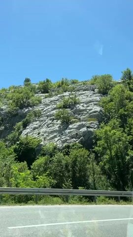 The video shows a view from a vehicle moving along a road, with a steep, rocky mountainside covered in lush green trees on the right. The sky is clear and blue. The mountainside has visible rock formations and some areas are covered with netting, likely for rockfall protection. The road appears to be paved with white lines marking the lanes. The overall impression is of a scenic drive through a mountainous region on a sunny day.