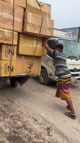 A man in a striped shirt and patterned skirt is pushing a small trailer loaded with cardboard boxes, which are secured with ropes. He is wearing sandals and is leaning heavily against the boxes to move the trailer, which is attached to a parked truck. The trailer and the truck are on a dirt road with some trash scattered around. An auto-rickshaw passes by. The boxes are labelled with "Zhen Patriotic Industries" and other text in English.