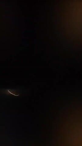 The video captures a nighttime scene over a body of water, likely the ocean. The sky is dark and overcast, with a bright, diffused light on the horizon, suggesting distant lightning or a storm. The water is calm, reflecting the dim light. The camera appears to be at a high vantage point, possibly from an airplane. The footage is grainy and somewhat blurred, indicating low light conditions and potentially movement. There are no people or distinct objects visible.