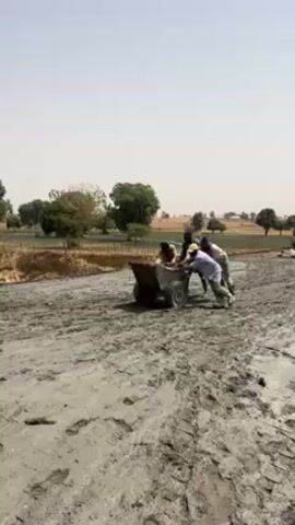 The video shows four men pushing a small concrete transport cart along a muddy path. Once they arrive at their destination, a man in rubber boots and a striped shirt starts to use a hoe to spread concrete over rebar.  The backdrop is a rural landscape with trees and a clear sky.