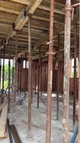 The video shows the interior of a building under construction. Multiple adjustable metal support posts, known as shoring poles, are visible, holding up wooden planks that form a temporary ceiling or floor structure. These shoring poles are made of rusty metal and are placed in a grid pattern throughout the space. The floor is concrete, and there are stacks of lumber and rebar on the ground, indicating ongoing construction work. The walls appear to be made of brick. In the background, there are glimpses of the outside, with trees and a roofline suggesting a residential or commercial building site. The scene is utilitarian and shows the structural support system for a construction project.