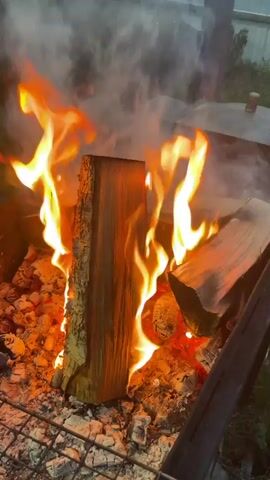 The video shows a close-up view of a burning fire in a fire pit, with thick logs fueling large flames that produce smoke.  The fire is burning on a metal grate within the pit. The flames are bright orange and yellow, rising from the burning logs and embers. The background is somewhat blurred.  The music playing is a fast-paced, electronic style.