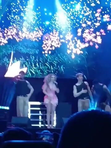 This video captures a live performance by a female singer on a concert stage. She's center stage, dressed in a short, pink floral outfit, and holding a microphone. Flanking her on both sides are male dancers in black tank tops and khaki pants, performing synchronized choreography. The background screen displays changing, vibrant graphics, including abstract shapes and a large, orange flower. The camera is at audience level, and you can see the heads and hands of the crowd in the foreground. The lighting is bright and colorful, typical of a concert setting.