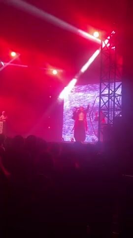 A concert performance is taking place on a stage. The stage is lit with pink and white lights, with beams of white light cutting across the scene from above. A large screen behind the performer displays abstract imagery. The performer, wearing a dark outfit with red fringe and a lighter colored shirt, is holding a microphone and gesturing with their free hand. They appear to be singing or speaking to the audience. There is smoke or fog on stage. The lower part of the frame shows the silhouetted heads and shoulders of an audience. Another person, also in a dark outfit with fringe, is visible to the right of the main performer, behind them. The scene is framed by a black metal truss structure on the right side of the image.
