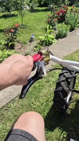 A first-person perspective video shows a person's hand gripping the handlebars of a red and white tiller in a garden. The tiller has large black tires and a red engine. In the background, there is a pathway lined with red roses and other green plants, leading to a white house. The sun is shining, casting shadows on the grass.