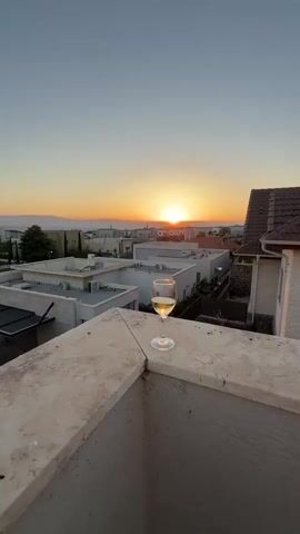 A glass of white wine sits on a textured, light-colored railing, overlooking a residential neighborhood as the sun sets. The sky is a gradient of pale blue at the top, transitioning to a warm orange and yellow near the horizon, where the sun is a bright, dominant orb. Silhouetted mountains are visible in the distance.

Below the railing, several modern, white-toned houses with flat roofs and air conditioning units are visible. Some have small gardens or courtyards. The setting appears to be a rooftop or balcony, offering a panoramic view of the suburban landscape. The time of day is clearly sunset, indicated by the warm, low light and the colorful sky. The overall mood is serene and peaceful, suggesting a moment of quiet enjoyment or relaxation.

The scene is set in Katzrin, Israel, a town in the Golan Heights. The architecture and landscape are consistent with this location. There is no visible text in the media.