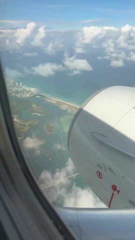 The view from an airplane window shows a city with tall buildings along a coastline, with an ocean next to the beach. There are islands with a lot of boats anchored around them. The sky is blue with white puffy clouds. Part of the airplane’s engine is visible in the foreground.