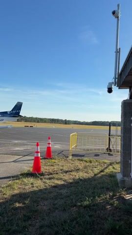 The video begins with a view of an airport tarmac on a clear day with a blue sky. A small airplane is visible in the background, with its tail number partially obscured. In the foreground, two orange traffic cones are placed on the grass next to a yellow barrier. A security camera is mounted on a pole attached to a building. The video then zooms in on the tarmac where a white jet airplane is taxiing. The airplane has a sleek design and appears to be a private jet. Trees and foliage line the background. The camera continues to track the jet as it moves across the tarmac.