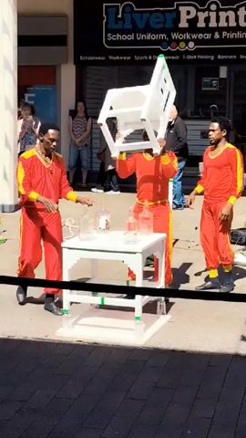 Three men are performing an outdoor street performance in front of a shop. Two men in red and yellow tracksuits are involved in balancing a white chair on top of a small white table. One man, on the left, is crouched by the table and seems to be adjusting something. The other man, on the right, is standing and holding the chair steady. A third man, also in a red and yellow tracksuit, is positioned behind the table, inside the chair, seemingly supporting it from within. The table has several glass bottles and jars on it. In the background, several bystanders are watching the performance. The shop behind them has a sign that reads "Liver Print School Uniform, Workwear & Printing".