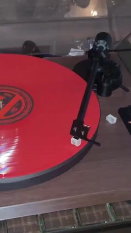 A close-up, slightly angled shot shows a red vinyl record spinning on a turntable. The turntable has a wooden base and a black tonearm with a white stylus that is in contact with the record, suggesting the music is playing. There is a partial view of the record's label, which appears to be black and orange with a stylized logo. To the right of the turntable, there are a couple of small white objects, possibly record accessories, and a portion of a black remote control. The background is softly blurred, showing a glass surface and what appears to be a dimly lit room. The lighting creates some specular highlights on the spinning record.