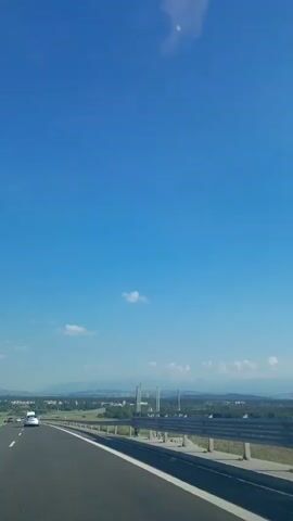 This is a vertical video recording of a scenic drive on a sunny day, likely on a highway outside Nowy Targ, Poland. The camera is positioned from the passenger seat of a vehicle, capturing the road ahead and the expansive blue sky above.

**Subjects:**
The primary subjects are the road, the surrounding landscape, and the sky. There are several vehicles visible on the road, including cars and a white van. The landscape consists of rolling hills, trees, and distant buildings or a town. The sky is a brilliant blue with a few scattered white clouds, one of which has a wispy, elongated appearance.

**Activity/Event:**
The video captures the experience of traveling on a highway. The movement of the vehicle is evident, indicated by the slight blur of the passing landscape and the dynamic perspective of the road. The overall impression is one of a journey through a picturesque area.

**Notable Details:**
*   **Time of Day:** The bright sunlight and clear blue sky suggest it is daytime, likely mid-morning to afternoon.
*   **Weather:** The weather appears to be very pleasant, characterized by clear skies and abundant sunshine.
*   **Location Cues:** While not explicitly stated in the visual, the mention of Nowy Targ, Poland provides the geographical context. The landscape suggests a rural or semi-rural setting with hills, which aligns with the topography of the region.
*   **Emotions:** The video evokes feelings of freedom, openness, and the enjoyment of a road trip. The bright colors and wide-open spaces contribute to a positive and relaxed mood.
*   **Visible Text:** No visible text is present in the video.