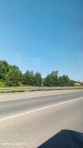 The video shows a view from a moving vehicle on a sunny day. The road is paved with a white line dividing the lanes. On the left side of the road, there are evergreen trees and houses with various colored roofs. There are also streetlights and power lines visible. The sky is clear and blue. The bottom left corner of the video displays "Infinix HOT 50".