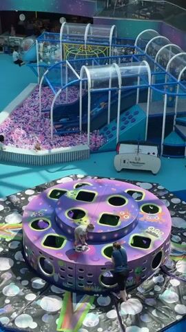 This media file captures a vibrant indoor playground, likely a children's entertainment center, with a cosmic or space theme. The main subjects are several people, including children actively playing and adults supervising.

The scene is dominated by a large, multi-level play structure featuring blue and purple netting, slides, climbing elements, and tubes. A prominent ball pit filled with pink and purple balls is a central feature. In the foreground, a circular, multi-tiered play structure with purple and green accents, decorated with cartoon alien characters and space-themed graphics, is also active with children and adults.

Notable details include the bright, artificial lighting, suggesting an indoor setting during daytime. The overall atmosphere appears lively and cheerful, with the children engaged in energetic play. The setting appears to be a large, open indoor space, possibly a mall or a dedicated entertainment complex, given the scale and design of the playground.

Visible text includes "TelkaBoom" on a white cart in the middle of the scene, and a sign on the play structure that also reads "TelkaBoom". Other text in the background, in Russian, is partially visible on escalators and signage, hinting at a location in Russia. The context provided indicates this is in Sovkhoza imeni Lenina, Russia.