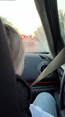 The video is shot from the perspective of someone inside a car, looking over the shoulder of another person. This person has short, light-colored hair and is wearing a black hoodie and light-colored pants. They are seated and wearing a seatbelt. The view outside the car shows a road with other vehicles ahead. The sun is setting, casting a warm glow and creating a lens flare effect. The interior of the car is visible, including the dashboard, air vents, and a red ambient light strip. The overall atmosphere appears to be that of a relaxed journey.