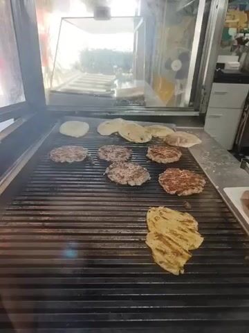 The video shows a close-up view of a flat-top grill with several items cooking on it. In the background, there is a reflection of what appears to be a street scene. On the grill, there are eight hamburger patties and some flatbreads. One piece of flatbread in the foreground has grill marks. The grill itself is made of black metal with parallel horizontal grates. The overall environment appears to be a commercial kitchen or food stand. The lighting is bright, likely from overhead lights or sunlight.