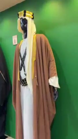 The video shows two mannequins displayed against a bright green wall. The mannequin on the right is dressed in a traditional Arabian outfit consisting of a white dishdasha, a brown bisht, and a keffiyeh with an agal. The outfit also includes a black leather belt with metal studs and crossed straps. To the left, the second mannequin wears a black abaya with colorful embroidery around the collar and hood. The abaya has a hood that is pulled up, concealing the mannequin's face with a geometric patterned covering. A small framed sign is visible on the wall above each mannequin. The camera focuses on each of the mannequins individually, moving from right to left.