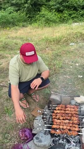 A group of people are having a barbecue outdoors. The camera pans across the scene, starting with a man in a red hat crouching by a grill with skewers of meat cooking over coals. He is wearing a green and white checkered shirt and blue shorts. The second shot focuses on two men sitting on the grass. One is wearing a green t-shirt and olive pants and is holding a small cup. The other is wearing a maroon t-shirt and dark shorts and is pointing. The third shot shows a man in a white t-shirt with glasses looking at the camera. Behind him, the man in the maroon t-shirt is still visible. The final shot shows a woman with her eyes closed and a smile on her face, holding up her hand with her fingers in a gesture. Behind her, a bridge is visible in the distance. The environment is grassy with trees and foliage in the background, suggesting a natural setting.