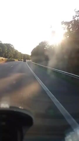 The video shows a view from inside a vehicle driving on a highway. The sun is low in the sky, creating lens flare and glare on the windshield. Trees line the right side of the highway, and the road curves slightly to the left. There are other vehicles visible in the distance.
