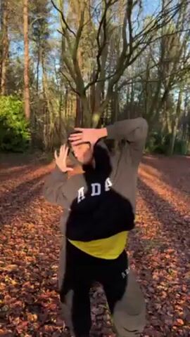 A woman is standing in a forest covered with dead leaves, wearing a black and grey tracksuit with the letters "ADA" in white. She pulls the hood over her head and walks along a path covered in leaves. She seems to be posing and showing off her tracksuit to the camera. The sun is shining through the trees. She says it's the first tracksuit she has ever owned.
