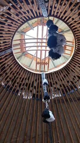 The video shows the interior of a yurt, looking upwards towards the central opening (tuunduk). The wooden lattice structure of the yurt walls and roof beams is visible, radiating outwards from the center. Decorative elements, including fabric bands with geometric patterns and hanging tassels made of yarn in black, white, and gray, are attached to the structure. The tuunduk is open, allowing sunlight to filter into the yurt, illuminating the wooden beams and the hanging decorations. The camera slowly pans upward, focusing on the tuunduk and the descending tassels.