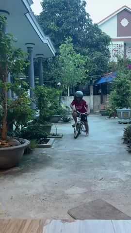 A child wearing a red shirt, black shorts, and a green helmet rides a bicycle on a paved outdoor patio. The child is in the center of the frame, looking towards the camera with a determined expression. The bicycle is small, suitable for a child. The patio is surrounded by lush green plants in large pots, and a building with a white and brown facade is visible in the background. The overall atmosphere appears to be a quiet, domestic setting, possibly a backyard or courtyard.