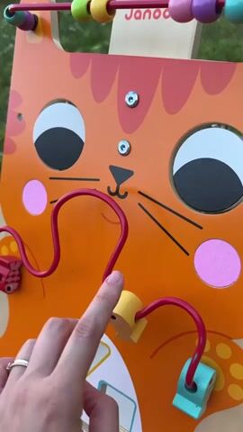 This is a close-up video of a child's wooden toy. The toy is an orange cat face with large, round black and white eyes, pink circular cheeks, and black whiskers. A red wire bead maze with yellow and blue beads, as well as small fish-shaped beads, is attached to the toy. A person's hand, wearing a ring, is interacting with the toy, moving a bead along the maze. The toy appears to be made of wood, with some painted details. There is a visible wooden abacus-like structure at the top of the toy with colorful beads. The background is blurred and suggests an outdoor setting, possibly a garden or grassy area. The lighting indicates it is daytime. The toy is branded with the text "Janod" visible on a wooden piece at the top. The activity is a child playing with a bead maze toy.