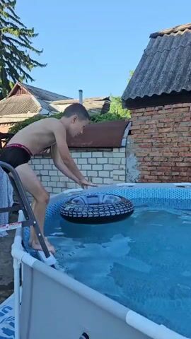 A young boy jumps into a pool with an inflatable tube. He lands in the water with a splash and then emerges, swimming. There is a red and white inflatable boat in the pool with him. The pool is in a backyard with a brick wall and a tiled roof in the background.