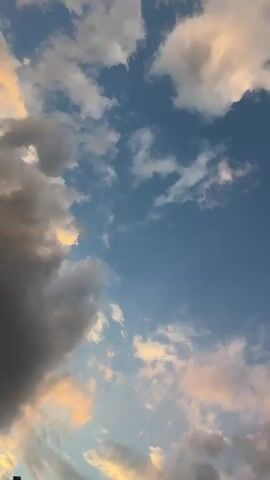 This is a vertical, low-angle shot of a sky filled with clouds. The sky is a clear, deep blue, with various types of clouds scattered across it. On the left side of the frame, there are large, dark, and imposing storm clouds. These clouds are casting shadows and appear to be quite dense. To the right of these darker clouds, the sky opens up to reveal lighter, wispy clouds that are tinged with hues of orange and pink, suggesting either sunrise or sunset. These lighter clouds are more spread out and allow more of the blue sky to be visible. The overall impression is one of a dynamic sky, possibly transitioning from or towards stormy weather.

There are no people or man-made objects clearly visible, except for a dark, indistinct shape at the very bottom left corner of the image, which might be the edge of a roof or building. The setting appears to be outdoors, with the focus entirely on the sky. The presence of both dark storm clouds and the warm, colored light on the other clouds hints at a time of day when light conditions are dramatic, likely early morning or late evening. The weather appears to be unsettled, with the possibility of rain indicated by the dark clouds, but also the potential for beautiful atmospheric light. The general feeling evoked is one of awe at the natural beauty and power of the sky, perhaps with a touch of melancholy or anticipation due to the contrasting cloud formations. No visible text is present in the media file.