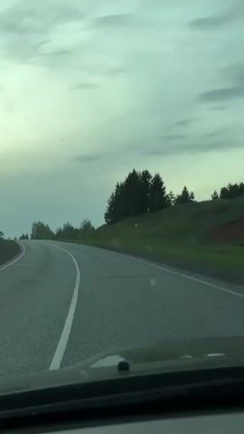 The video depicts a drive along an asphalt road, likely in a rural or semi-rural area. The road has white painted lines and appears to be somewhat damp or wet. The surrounding environment consists of grassy areas and patches of forest or clusters of trees. An embankment with visible soil erosion is visible. The sky is overcast, with the sun intermittently peeking through the clouds. A tall telecommunications tower appears in the distance. The footage is shot from the driver's perspective, offering a view through the car's windshield, including part of the dashboard.