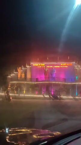 The video shows a nighttime view from inside a vehicle, looking out at a street scene. The street is decorated with lights, and there are statues and illuminated structures visible. The primary focus appears to be a large, illuminated monument with statues of figures. There are vehicles on the road, including motorcycles and cars. The lighting is predominantly artificial, with bright lights and reflections creating a hazy effect. The overall atmosphere suggests a public space or an event being celebrated at night.