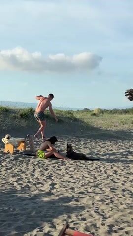 A sunny beach scene features a man attempting to balance on a small wooden stool. He's barefoot, shirtless, and wears patterned swim trunks. A woman sits on the sand, wearing a yellow bikini top and green bottoms, looking at her phone. A German Shepherd dog lies next to her. The man is precariously balancing with his arms out, seemingly struggling to maintain his balance. A few wooden stools and a towel are nearby. The background includes grassy dunes, mountains, and a partially visible thatched structure. The sky is blue with a few white clouds.
