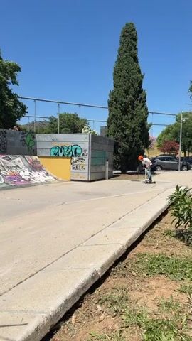 A person wearing a white t-shirt, blue jeans, and an orange helmet rides a silver scooter at an outdoor skatepark in Esplugues de Llobregat, Spain, on a bright, sunny day. The individual approaches a graffiti-covered quarter pipe, rides up the ramp, and performs a stall trick on the coping. The scooter and rider briefly pause on the edge of the ramp before dropping back down. The person then rides away, smiling after successfully completing the maneuver.

The skatepark features concrete ramps and walls, extensively covered in colorful graffiti with discernible tags like "MILO," "ERIK," "NIKO," "LUXITO," "FESH," "FOREVER," "SOH," and "HOOD." In the background, green trees line the area, and hills are visible under a clear blue sky, with what appears to be a tower on a distant hilltop. A metal fence runs along the top of the skatepark walls. The ground is concrete, with a grassy area to the right.